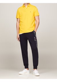 Man som bär en gul kortärmad polo, marinblå joggerbyxor med "Tommy Hilfiger"-text, och vita sneakers, stående mot en neutral bakgrund.