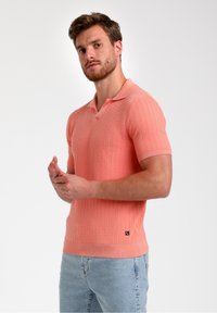 Světle korálová polokošile s krátkým rukávem, texturovanými svislými pruhy, klasickým límcem a žebrovaným lemem, spárovaná se světle modrými džíny.