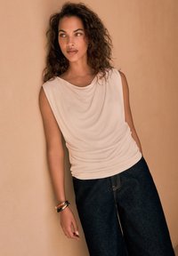 Top drappe beige chiaro con scollatura rotonda, abbinato a jeans larghi neri. La modella indossa braccialetti al polso. Texture del tessuto morbida.