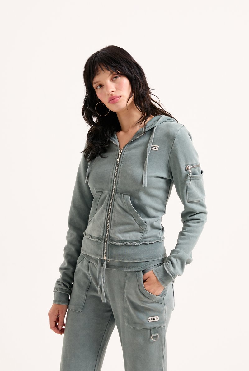 Vrouw in een vervaagde teal kleurige ritsjack en bijpassende sweatpants met zakken, staand met één hand in de zak tegen een witte achtergrond.