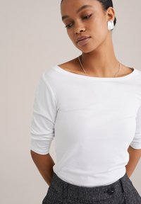 Frau mit dunkler Haut und zurückgepulltem Haar trägt ein weißes Langarmshirt, eine silberne Halskette, große silberne quadratische Ohringe und dunkle, geknöpfte Hosen.