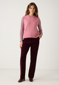 Helles rosa ärmelloses Stricktop über einer gemusterten langärmeligen Bluse, kombiniert mit dunkelburgunderfarbenen Cordhosen und braunen Schuhen.