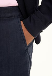 Pantaloni di lino blu navy con tasca laterale, abbinati a una camicia scura. Il tessuto appare leggermente strutturato e traspirante.