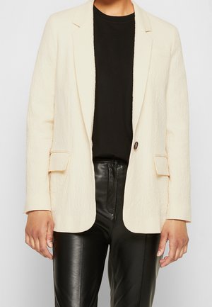 Personne portant un blazer texturé crème sur un haut noir assorti à un pantalon en cuir noir, montré des épaules aux hanches.