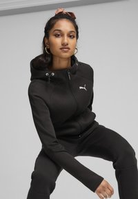 Sudadera deportiva negra con cremallera en el frente, capucha con cordón y logotipo. Diseño ajustado con textura suave y acentos contrastantes en las mangas.