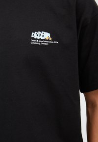 Svart bomull t-shirt med en liten grafisk logotyp på vänster bröst med text och stjärnor. Korta ärmar och en klassisk rundhalsdesign.