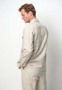 Camicia di lino beige con vestibilità rilassata, maniche lunghe e polsini con bottoni. Presenta una singola tasca a soffietto sul lato sinistro del petto.