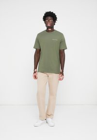 T-shirt en coton vert olive à manches courtes, col rond et petit logo blanc. Associé à un pantalon droit beige clair et des baskets blanches.