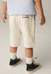 Bambino che indossa pantaloni corti crema e scarpe alte nere con calzini bianchi, visto da dietro su sfondo neutro.