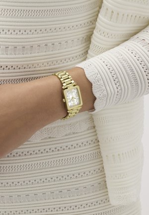 Zlatni pravokutni ručni sat s rimskim brojkama na osobi koja nosi bijelu teksturiranu majicu dugih rukava.