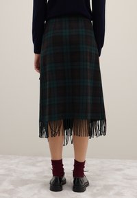 Jupe écossaise en vert foncé et marron, avec un ourlet à franges. Portée avec des chaussettes bordeaux et des chaussures noires. Tissu en mélange de laine lisse.