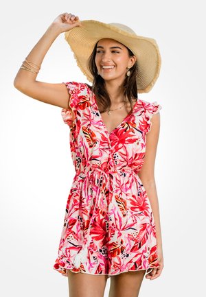 Donna che sorride, indossa un cappello di paglia a tesa larga e un vestito senza maniche a fiori rosa, tenendo la tesa del cappello con una mano.
