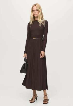 Donna con lunghi capelli biondi, che indossa un vestito plissettato a maniche lunghe di colore marrone scuro, tacchi neri con cinturini e tiene in mano una borsa nera.