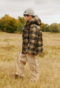 Padded jacket in gray, black, and brown check pattern, with a hood. Beige cargo pants. Knit gray beanie. Standing in a grassy field.