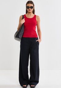 Červený žebrovaný top bez rukávů, černé široké kalhoty a černá taška. Modelka nosí černé oversized sluneční brýle a černé šlapky. Hladké textury.