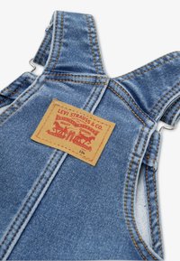 Salopetă din denim albastru cu cusături galbene, prevăzută cu bretele ajustabile pentru umeri și o etichetă de piele căprui cu grafice roșii și negre. Mărimea 12 luni.