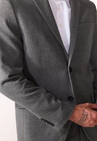 Grå teksturert blazer med notched lapels, enkel knappelukking og fire knappene på mansjettene. Bæres over en hvit skjorte med en synlig lomme.