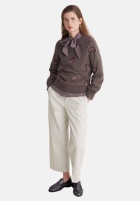 Maglione marrone lavorato a maglia con ricamo floreale, abbinato a pantaloni larghi crema e mocassini neri. Presenta un colletto con fiocco.