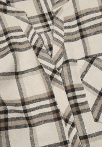 Beige geruite stof met zwarte en bruine lijnen, met een zachte textuur, die overlappende patronen en een gedeeltelijke kraag bij de hals toont.