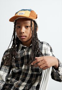 Casquette en velours côtelé marron, bleu et jaune avec étiquette, dotée d'une douce doublure blanche. Chemise en flanelle à carreaux noir et blanc avec des boutons marron.
