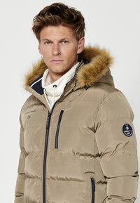 Įdegus puffer tipo striukė su dirbtinio kailio pamušalu ant gobtuvo, užtrauktuku priekyje ir šoninėmis kišenėmis. Ant kairiojo rankovės yra logotipo užrašas, o audinys yra tekstūruotas.