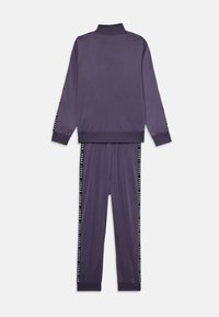 Lilla tracksuit laget av glatt stoff, med jakke med høy krage og bukser med svarte og hvite sidelister. Myke, tettsittende mansjetter og liv.