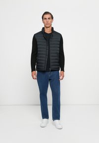Young man standing, wearing a black quilted sleeveless jacket over a black long-sleeve shirt, blue jeans, and white sneakers on a white background.