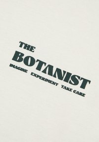 Witte stof met de tekst "THE BOTANIST" in donkergroene vette letters, met daaronder kleinere tekst "IMAGINE EXPERIMENT TAKE CARE".