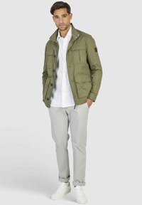 Veste vert olive avec plusieurs poches frontales, fermeture à boutons et col montant, assortie à un pantalon gris clair et des baskets blanches.