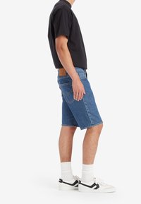 Man som bär svart t-shirt, blå denimshorts, vita strumpor och vita sneakers med svart rand, står på sidan mot vit bakgrund.