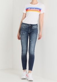 Camiseta blanca con el texto "Rainbow" en varios colores, combinada con jeans ajustados azules con acentos desgastados y zapatillas blancas con detalles en rosa.