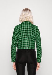 Vihreä ja musta houndstooth-kuvioitu lyhyt takki, jossa on terävöitetty kaulus ja pitkät hihat, valmistettu teksturoidusta kankaasta. Mustat slim-fit-housut näkyvissä.