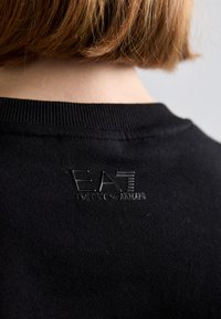 Μαύρο φούτερ με υφασμάτινο λαιμό, με ανάγλυφο λογότυπο "EA7" κεντημένο στο πίσω μέρος. Μαλακό βαμβακερό ύφασμα με λεία υφή.