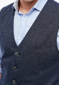 Marineblaues Tweed-Weste mit strukturiertem Design, V-Ausschnitt, fünf Knöpfen und einer Brusttasche. Über einem hellblauen Hemd getragen.