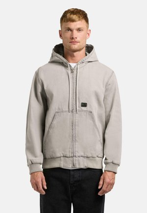 Mand iført lysegrå hættetrøje med lynlås og forlommer samt sorte bukser, stående foran en ensfarvet hvid baggrund.