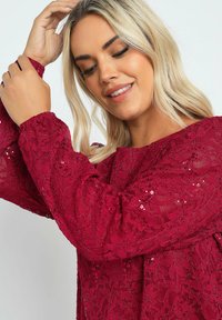 Abito in pizzo bordeaux con maniche lunghe e voluminose. Il tessuto presenta motivi floreali e paillettes incorporate. Vestibilità leggermente svasata e texture morbida.