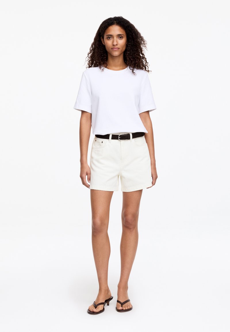 Femme aux cheveux bouclés portant un t-shirt blanc uni, un short crème avec une ceinture noire et des sandales à talons noires, debout sur un fond blanc.