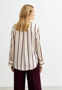 Langsleeve blouse in crèmekleur met verticale kastanjebruine strepen. Heeft een losse pasvorm, knoopmanchetten en een gebogen zoom aan de achterkant.