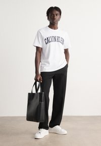 Hvit bomull t-skjorte med "CALVIN KLEIN" trykket i marineblå, kombinert med svarte bukser, holder en svart lær tote bag og har på seg hvite sneakers.