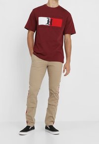 Maroon short-sleeve t-shirt featuring a white and red logo block, paired with beige pants that have red side stripes and black sneakers.