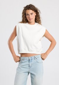 Mujer con cabello rizado, vistiendo un top corto sin mangas blanco y jeans de tiro alto de color azul claro, con las manos sujetando la cinturilla.