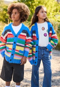 Colorful knitted cardigans with stripes in blue, red, green, and yellow, featuring large buttons. One wears a white shirt with smiley face patches.