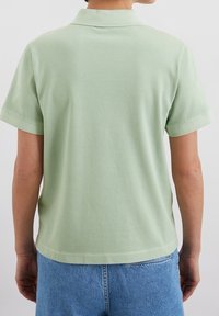 Henkilö, jolla on vaaleanvihreä lyhythihainen polo paita ja siniset farkut, kuvattuna selkäpuolelta yksinkertaista valkoista taustaa vasten.