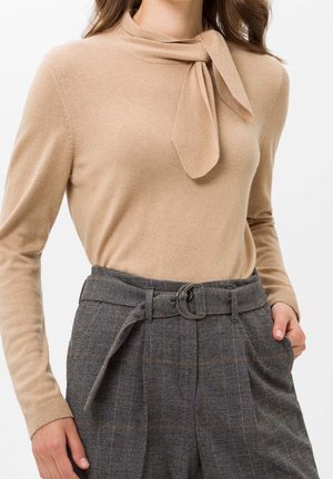 Kobieta ubrana w beżową bluzkę z długim rękawem i wiązaniem przy szyi oraz szare spodnie w kratę z wysokim stanem, paskiem i okrągłą klamrą, z ręką w kieszeni.