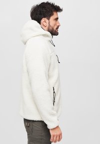 Pullover bianco in pile con cappuccio, dotato di cordini neri regolabili. Texture morbida e vestibilità comoda, con tasche laterali.