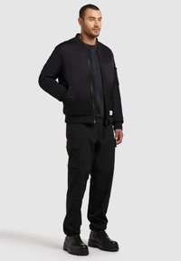 Giubbotto bomber nero con zip frontale, polsini a costine e tasche laterali, abbinato a pantaloni cargo neri e stivali neri stringati.