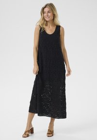 Black sleeveless dress featuring intricate circular lace patterns, longer length, and a relaxed fit, paired with brown block-heeled sandals.