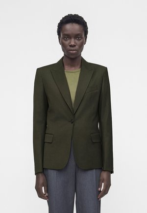 Femme aux cheveux courts et bouclés portant un blazer vert olive sur un haut vert clair et un pantalon gris, debout devant un fond blanc uni.