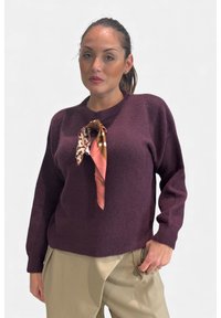 Maglione lavorato a maglia color bordeaux con maniche lunghe, scollo rotondo e una sciarpa di seta con motivo annodata davanti, indossato con pantaloni beige.