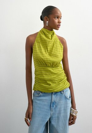 Femme portant un haut halter vert à carreaux sans manches et un jean bleu clair, regardant sur le côté, avec des boucles d'oreilles argentées et plusieurs bracelets.